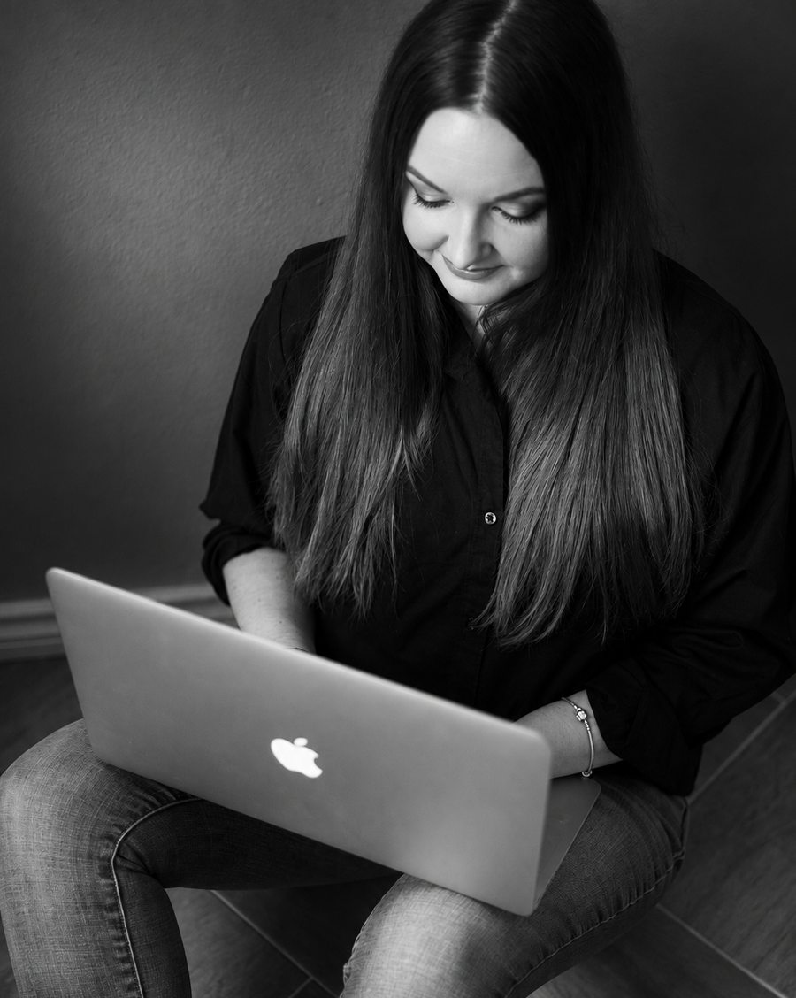 A woman with long dark hair sits on the ground using a silver Apple laptop. The photo is in black and white, showing her in a black button-up shirt and jeans against a dark gray wall and wood floor.
